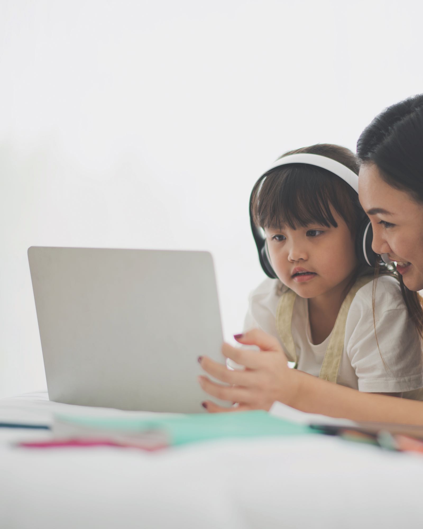 Young Asian mother and daughter, reading homework on computer laptop on white bed at home smiling and feeling happy.